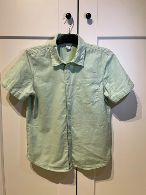 Ljusgrön kortärmad linneskjorta - Snygg ljusgrön skjorta i linne med korta ärmar och klassisk krage. Skjortan har bröstficka och knäppning framtill. Perfekt för varma dagar och enkel att styla till olika outfits.