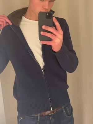 Mörkblå cardigan jacka med fuskpäls - Mörkblå cardigan jacka, med dragkedja och mjuk brun fuskpäls på kragen. Jackan är stickad och har långa ärmar samt ribbade muddar. Perfekt för dig som vill ha en stilren och mysig look.   Jag kan skicka mer bilder också,Och du kan ta av pälsen och sätta på den igen