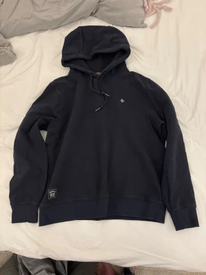 Mörkblå hoodie från Morris - Mörkblå hoodie från Morris med liten broderad logga på bröstet och justerbar dragsko i huvan. Mjuk bomullskänsla och ribbade muddar. Perfekt för en chill och stilren look.