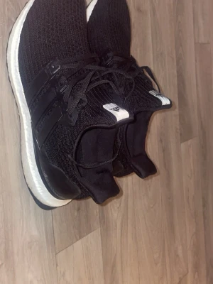 Svarta Adidas Ultraboost sneakers - Säljer ett par svarta Adidas Ultraboost sneakers med stickad ovandel och klassiska tre ränder på sidan. Skorna har snörning, platt sula och vit mellansula som ger skön dämpning. Perfekta för dig som gillar stilrena och bekväma sneakers. Knappt använda 