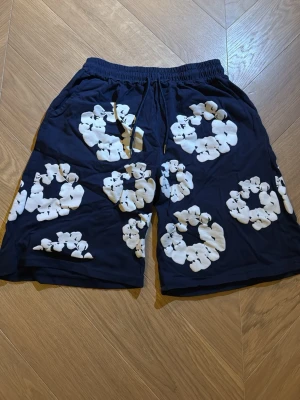 Mörkblå shorts med blommigt mönster - Snygga mörkblå shorts med vita blommiga tryck och elastisk midja med snörning. Perfekta för sommardagar och chill häng. Shortsen har sidofickor och en bakficka, samt en loose fit för extra komfort.