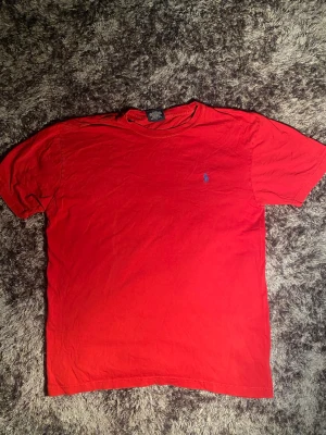 Röd t-shirt från Polo Ralph Lauren - Säljer en röd t-shirt från Polo Ralph Lauren med klassisk blå broderad logga på bröstet. T-shirten har rund hals och korta ärmar, tillverkad i mjuk bomull. Perfekt för dig som gillar stilrena och ikoniska plagg.