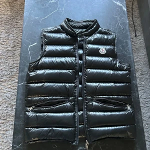 Moncler väst - Snygg svart dunväst från Moncler med glansig finish och klassisk logga på bröstet. Västen har ståkrage, två dragkedjefickor och quiltad design. Perfekt att slänga över en hoodie eller tröja för extra värme och stilpoäng. Äktehetsbevis på bild 4 och 5. Monclers storlek 1 sitter som xs/s.