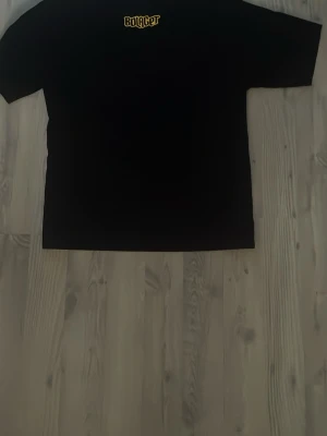 Svart Bolaget Rodeo t-shirt - Svart t-shirt från Bolaget Rodeo med stor tryckt logga i gult och lila på ryggen och mindre logga framtill. Klassisk rund hals och korta ärmar. Materialet känns som bomull, perfekt för dig som gillar streetwear med tydlig grafisk stil. Väldigt stor till storleken.
