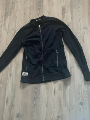 Marinblå kofta från Sail Racing - Marinblå kofta från Sail Racing med dragkedja framtill och två fickor med dragkedja. Jackan har en stilren look med ribbade muddar och diskret logotyp på nedre kanten. Perfekt för dig som gillar sportig och clean stil.