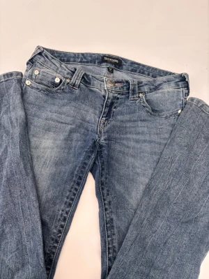 True Religion blå jeans, storlek 27 - Säljer ett par klassiska blå jeans från True Religion med ikonisk broderad ficka bak och silverfärgade knappar. Jeansen har en normal passform och raka ben, med ljusblå tvätt och kontrastsömmar i rosa. Materialet är slitstark bomull och jeans. Perfekt som julklapp. Köpt för 1400kr, pris kan diskuteras.