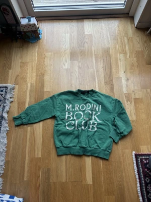 Grön sweatshirt från Mini Rodini - Säljer en grön sweatshirt från Mini Rodini med trycket 'M. Rodini Book Club' i vitt på framsidan. Tröjan har rund hals, ribbade muddar vid ärmslut och nederkant samt lång ärm. Perfekt för dig som gillar coola prints och avslappnad stil.