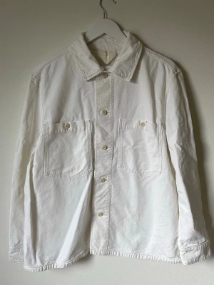 Vit overshirt från Arket i bomull - Säljer en stilren vit overshirt från Arket i 100% bomull. Jackan har klassisk krage, två stora bröstfickor med knappar och knäppning framtill. Perfekt att slänga på över en t-shirt för en clean look. Passar dig som gillar minimalistisk stil. 5M17