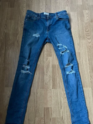 Blå skinny jeans från Jack & Jones - Säljer ett par blå skinny fit jeans från Jack & Jones, modell Liam. Jeansen har slitna detaljer och hål på både fram- och baksida, klassisk femficksdesign och bruna sömmar. Materialet är jeans med en snygg tvättad look och låg midja.