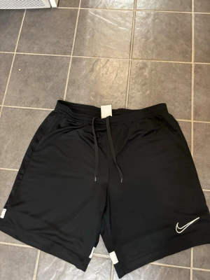 Svarta träningsshorts från Nike - Säljer ett par svarta träningsshorts från Nike med vit broderad logga på benet. Shortsen har snörning i midjan och är gjorda i mjukt och lätt polyester-material. Perfekta för gymmet eller sport. Klassisk loose passform och stilren design.