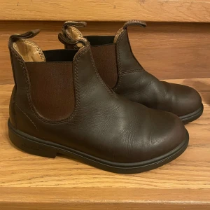 Blundstobe bruna boots i skinn Stlk 2 (35) - Säljer ett par klassiska Chelsea boots från Blundstone i mörkbrunt skinn med elastiska sidopaneler och rund tå. Skorna har dragflikar både fram och bak för enkel påtagning. Perfekta för dig som gillar stilrena och tidlösa boots.