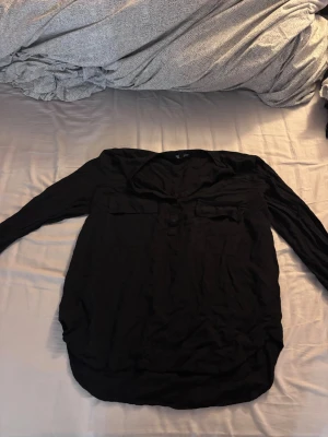 Svart skjorta - Svart skjorta från KappAhl i storlek M. Skjortan har lång ärm, två bröstfickor med lock och knappar framtill. Materialet känns lätt och mjukt, perfekt för en chill och stilren look. Passformen är normal och skjortan har en klassisk rundad nederkant.