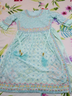 Ljusblå långklänning med broderi - Säljer en ljusblå långklänning med glittriga paljetter och broderade blommor i pastellfärger längs kanterna. Klänningen har trekvartsärm, snörning med tofsar i midjan och ett skirt, mönstrat tyg. Ingår matchande byxa och sjal i samma färg.