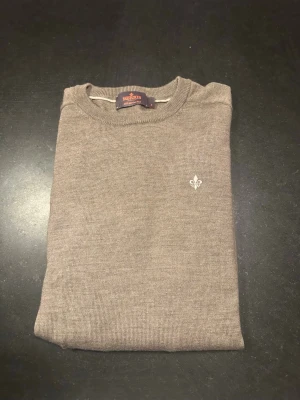 Beige stickad tröja från Morris - Säljer en beige långärmad stickad tröja från Morris med rund hals och diskret broderad logga på bröstet. Tröjan har en klassisk och stilren design som passar perfekt till jeans eller chinos. Perfekt för dig som gillar enkel och snygg stil.