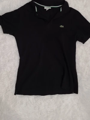 Svart piké från Lacoste, storlek M - Klassisk svart pikétröja från Lacoste med korta ärmar och krage med gröna och vita detaljer. Tröjan har den ikoniska krokodilloggan broderad på bröstet. Tillverkad i mjuk bomull som är skön mot huden och passar perfekt till jeans eller shorts.