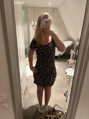 Svart blommig kortklänning - Säljer en svart kortklänning med rosa och gröna blommor. Klänningen har korta puffärmar,söt volangkant längst ner och har scrunch i mitten fram. Säljer för den inte använts någon gång!