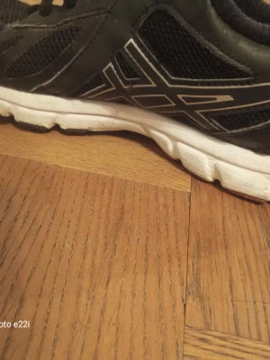 Svarta sneakers med vit sula - Säljer ett par svarta Asics sneakers med vit, grov sula och mesh-paneler på sidorna för extra ventilation. Skorna har snörning och en snygg silverfärgad logga på sidan. Perfekta för dig som gillar sportig stil och vill ha bekväma skor till vardagen.