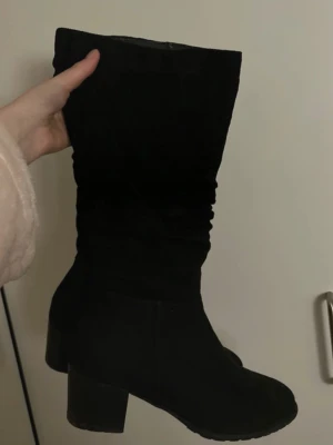 Svarta mockastövlar med klack - Säljer ett par svarta stövlar i mocka med rund tå och stabil klack. Stövlarna har en enkel och stilren design som passar till många olika outfits. Materialet är mjukt och ger en snygg look, perfekt för höst och vinter.