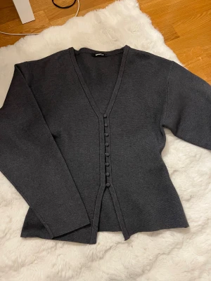 Mörkgrå kofta från Gina Tricot - Säljer en stilren mörkgrå kofta från Gina Tricot i storlek S. Koftan har v-ringning, långa ärmar och klädda knappar framtill. Materialet känns mjukt och är perfekt för lager på lager under höst och vinter. Så oanvänd knappt använt denna max 3 gånger 