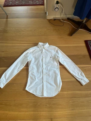 Vit slim fit skjorta från Ralph Lauren - Säljer en vit skjorta från Ralph Lauren i slim fit-modell, storlek XS men passar mycket bra på S. Skjortan har klassisk krage, långa ärmar och knappar framtill. Materialet är bomull och den har den ikoniska lilla broderade loggan på bröstet. Skjortan är använd fåtal gånger och saknar defekter och är perfekt för dig som gillar stilrena och tidlösa plagg. 