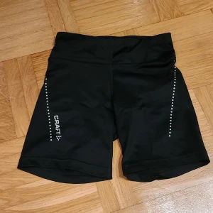 Svarta träningstights-shorts från Craft - Svarta träningstights-shorts från Craft i storlek S. De har en tight passform, hög midja och är gjorda i ett stretchigt syntetmaterial. Snygga detaljer med vita prickar längs sidorna och Craft-logga på ena benet. Perfekta för löpning eller gymmet.