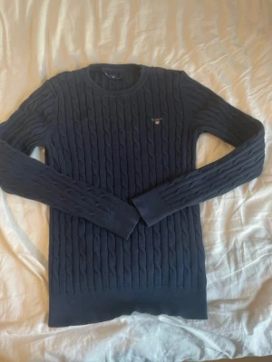 Mörkblå stickad tröja från GANT - Säljer en mörkblå stickad tröja från GANT i storlek S. Tröjan har ett klassiskt kabelstickat mönster, rund hals och långa ärmar. Diskret GANT-logo broderad på bröstet. Perfekt för dig som gillar stilrena och tidlösa plagg.