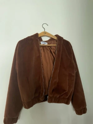 Brun teddyjacka med huva från Desired Fashion - Mysig brun teddyjacka med huva från Desired Fashion. Jackan har dragkedja framtill, elastiska muddar och är tillverkad i mjuk polyester. Perfekt för dig som gillar chill och comfy vibes. Fodrad insida för extra värme.