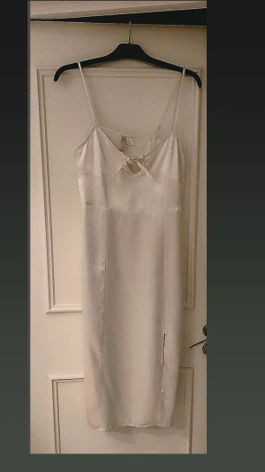 Vit satin klänning från H&M Divided - Säljer en vit långklänning i satin från H&M Divided. Klänningen har tunna axelband, en söt rosettdetalj framtill och en liten slits på sidan. Perfekt för sommarens alla tillfällen och ger en riktigt fräsch look.