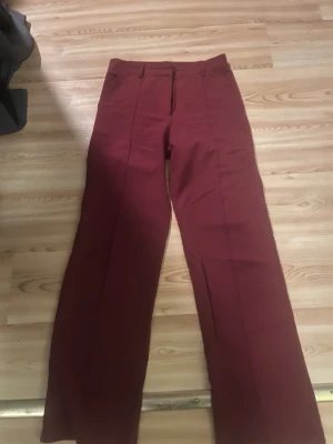 Mörkröda kostymbyxor - Snygga röda kostymbyxor med klassiska pressveck och rak passform. Byxorna har bälteshällor och är tillverkade i ett mjukt tyg som ger en stilren look. Perfekta för dig som vill ha en clean och modern vibe. Lite skadade på byxsluten pågrund att dem varit för långa på mig.