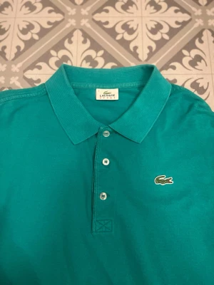 Turkos pikétröja från Lacoste - Säljer en klassisk turkos pikétröja från Lacoste Sport med krage och tre knappar framtill. Tröjan har korta ärmar och den ikoniska krokodilloggan broderad på bröstet. Perfekt för en chill och sportig look. Storlek 7/XL och Skicket är 9/10 och priset kan självklart diskuteras 🙂 tveka inte på att höra av dig om du funderar över något. /Isak