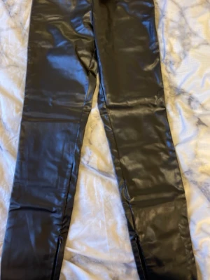 Svarta skinnbyxor från Bik Bok XS - Säljer ett par svarta skinnbyxor från Bik Bok i storlek XS. Byxorna har en glansig finish och är tighta med hög midja. Materialet är syntetiskt skinn och de har dragkedjor vid bensluten för extra stil. Perfekta för dig som vill ha en edgy look.