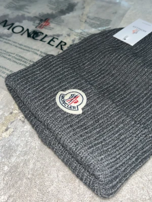 Grå Moncler stickad mössa - Snygg grå stickad mössa från Moncler med klassisk ribbad design och broderad logotyp framtill. Mössan har en bred uppvikt kant och är tillverkad i mjukt material som håller dig varm. Perfekt accessoar för kyliga dagar.
