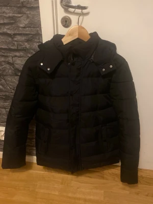 Svart pufferjacka med huva - Snygg och varm svart jacka med huva. Sparsamt använd och i mycket bra skick. Inga fläckar eller skador. Passar perfekt till höst/vinter och är lätt att matcha till olika outfits.