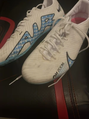 Nike Mercurial Zoom fotbollsskor 42.5 - Nike Mercurial Zoom fotbollsskor i vitt med blå och svarta detaljer samt rosa och svarta dobbar. Skorna har snörning och är tillverkade i syntetmaterial med mönstrad design på sidorna. Perfekt för dig som vill ha grymt grepp och lätt känsla på planen.