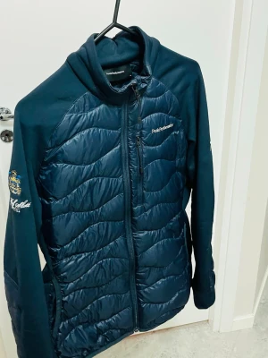 Mörkblå pufferjacka Peak Performance - Snygg mörkblå pufferjacka från Peak Performance med quiltad framsida och rygg, dragkedja hela vägen och hög krage. Jackan har långärmade partier i mjukt material och en bröstficka med dragkedja. Perfekt för dig som gillar stil och funktion.