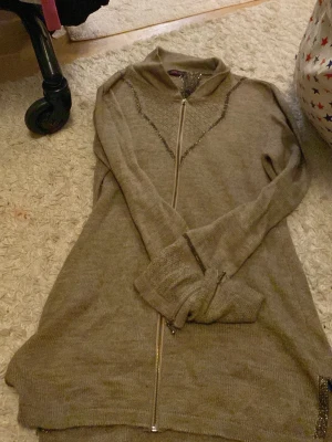 Beige lång kofta från Sincap - Lång beige kofta från Sincap med glittriga detaljer och hel dragkedja framtill. Stickad i mjukt material och har långärmad design. Snygga glittriga ränder längs axlar och ärmslut ger extra stil. Perfekt för lager-på-lager under kyliga dagar.