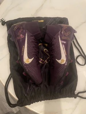 Nike Mercurial fotbollsskor lila/guld - Säljer ett par Nike Mercurial fotbollsskor i mörklila med guldiga detaljer och vit swoosh. Skorna har snörning, högt skaft och mönstrade grafiska inslag på sidan. Ovandelen är i syntetmaterial och designen är riktigt sportig och modern. Använts på en träning 