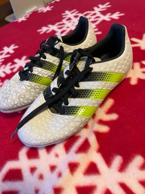 Adidas 16.3 vita fotbollsskor - Säljer ett par Adidas 16.3 fotbollsskor i vitt med svarta och neongula ränder. Skorna har snörning och en mönstrad ovandel i syntetmaterial. Adidas-logga i blått på hälen och insidan. Perfekta för dig som vill sticka ut på planen.