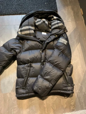 Burberry jacka  - Svart pufferjacka från Burberry med klassisk logotyp på bröstet och rutigt foder i huvan. Jackan har dragkedjor på fickorna och en quiltad design som ger extra volym. Perfekt för kalla dagar och har en riktigt snygg streetstil.