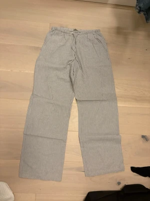 Randiga linnebyxor från Gina Tricot XS - Säljer ett par helt nya grå linnebyxor från Gina Tricot i storlek XS. De har använts endast ett fåtal gå gånger. Byxorna har vita vertikala ränder, elastisk midja med snörning och raka ben. Perfekta för chill dagar och har en skön loose passform.
