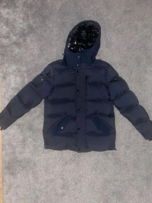 Mörkblå pufferjacka med huva - Säljer en mörkblå pufferjacka med avtagbar huva och dragkedja framtill. Jackan har stora fickor med lock och knappar, samt en extra ficka med dragkedja på ärmen. Insidan av huvan är glansig och jackan är perfekt för kalla dagar. Jackan är en perfekt fit om du är mellan M-L, original pris 10000