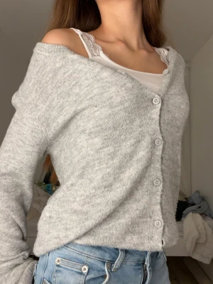 Ljusgrå stickad kofta med knappar - Mysig ljusgrå stickad kofta med knappar framtill. Koftan har lång ärm och en loose passform som ger en chill vibe. Perfekt att slänga över en topp eller linne. Enkel design utan mönster, passar till jeans eller kjol.