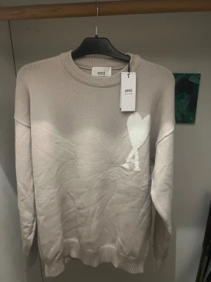 Beige stickad tröja från Ami - Säljer en beige stickad tröja från Ami Alexandre Mattiussi med ett stort vitt hjärta på bröstet. Tröjan har rund hals, ribbade muddar och lång ärm. Perfekt för dig som gillar stilrena detaljer och mjuka material.
