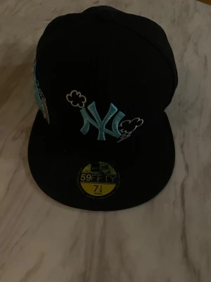 Svart New Era 59FIFTY NY keps - Säljer en svart New Era 59FIFTY keps med broderat NY Yankees-logo i ljusblått och vita molndetaljer. På sidan finns en broderad frihetsgudinna i blått och grått. Undersidan av skärmen är ljusblå. Perfekt för dig som gillar streetwear och snygga detaljer.