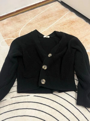Svart ribbad kofta med knappar - Säljer en svart ribbad kofta med tre stora knappar framtill. Koftan har en v-ringad design och är långärmad med en croppad passform. Perfekt att slänga på över en t-shirt eller linne för en chill look.