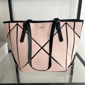 Rosa shoppingväska från Tosca Blu - Snygg shoppingväska i ljusrosa med svarta geometriska detaljer och svarta axelband. Väskan har ett modernt, stilrent utseende och är perfekt för dig som vill ha plats med allt du behöver. Tillverkad i syntetmaterial och har Tosca Blu-logga framtill.