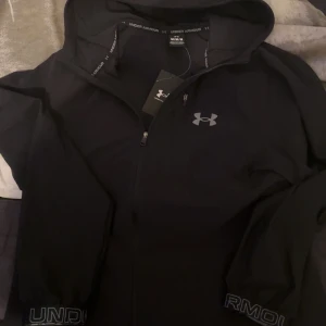 Svart vindjacka från Under Armour M - Svart vindjacka från Under Armour med huva och dragkedja framtill. Jackan har logga på bröstet och tryckt text på ärmsluten. Lätt och sportig modell i syntetmaterial, perfekt för aktiva dagar.