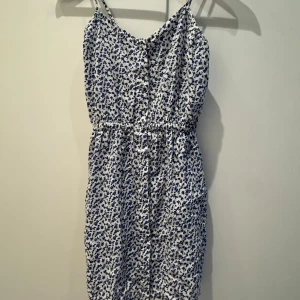 Blommig sommarklänning från JC - Supersöt kort klänning från JC i vitt med blått blommönster. Klänningen har smala axelband, knappar framtill och resår i midjan för en skön passform. 