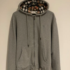 Grå tröja från Burberry - Grå hoodie från Burberry med klassiskt rutigt foder i huvan, broderad logga på bröstet och hel dragkedja framtill. Tröjan har snörning i huvan och två stora fickor. Perfekt för dig som gillar stilrena och exklusiva plagg. Knappt använd.