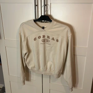 Beige sweatshirt från H&M Divided - Beige sweatshirt från H&M Divided med trycket 'COBRAS Arizona Cobras' i brunt framtill. Tröjan har rund halsringning, långa ärmar och ribbade muddar. Perfekt för en chill och avslappnad stil.